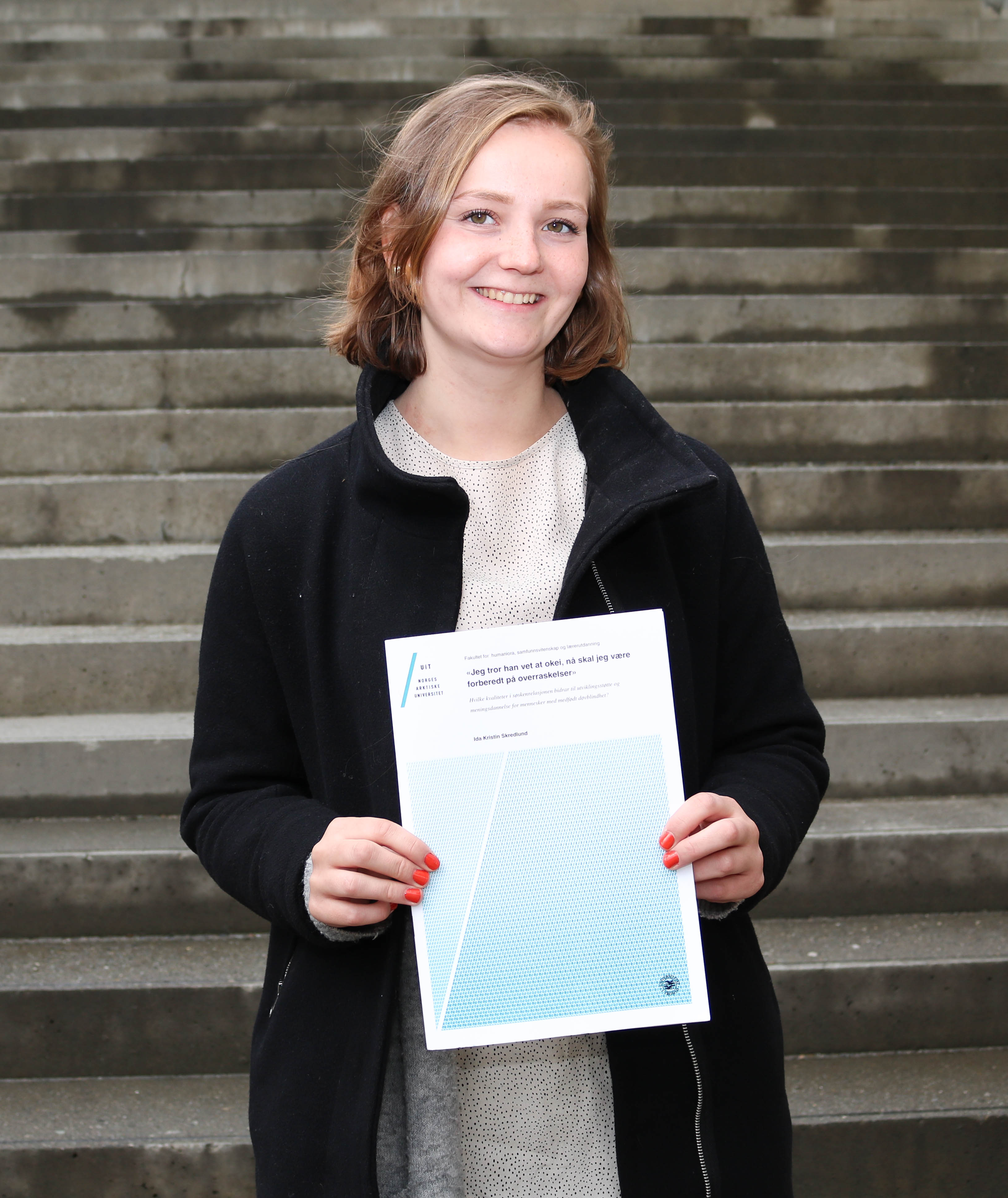 Ida Skredlund smiler og viser fram sin masteroppgave, stående foran betongtrappene ved Tromsø bibliotek og byarkiv.