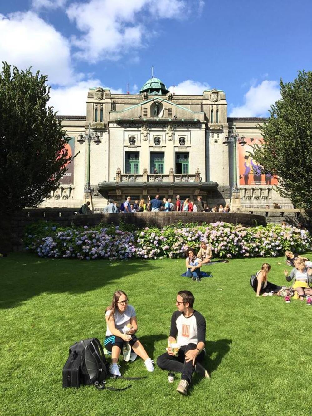 Den Nationale Scene i Bergen en solfylt lørdag, flere grupper med unge folk sitter i gresset foran teateret.