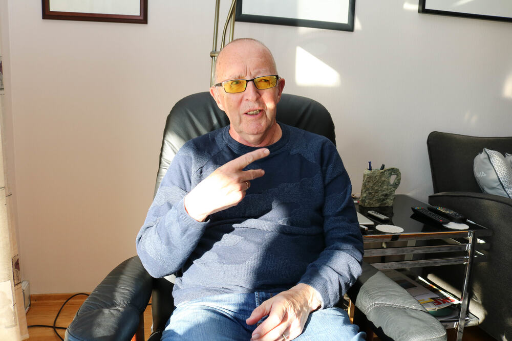 Eldre døvblind mann med gule fargefilterbriller sitter i sin godstol. Han viser med sin høyre hånd en v-håndform mot sofagruppen i stua.