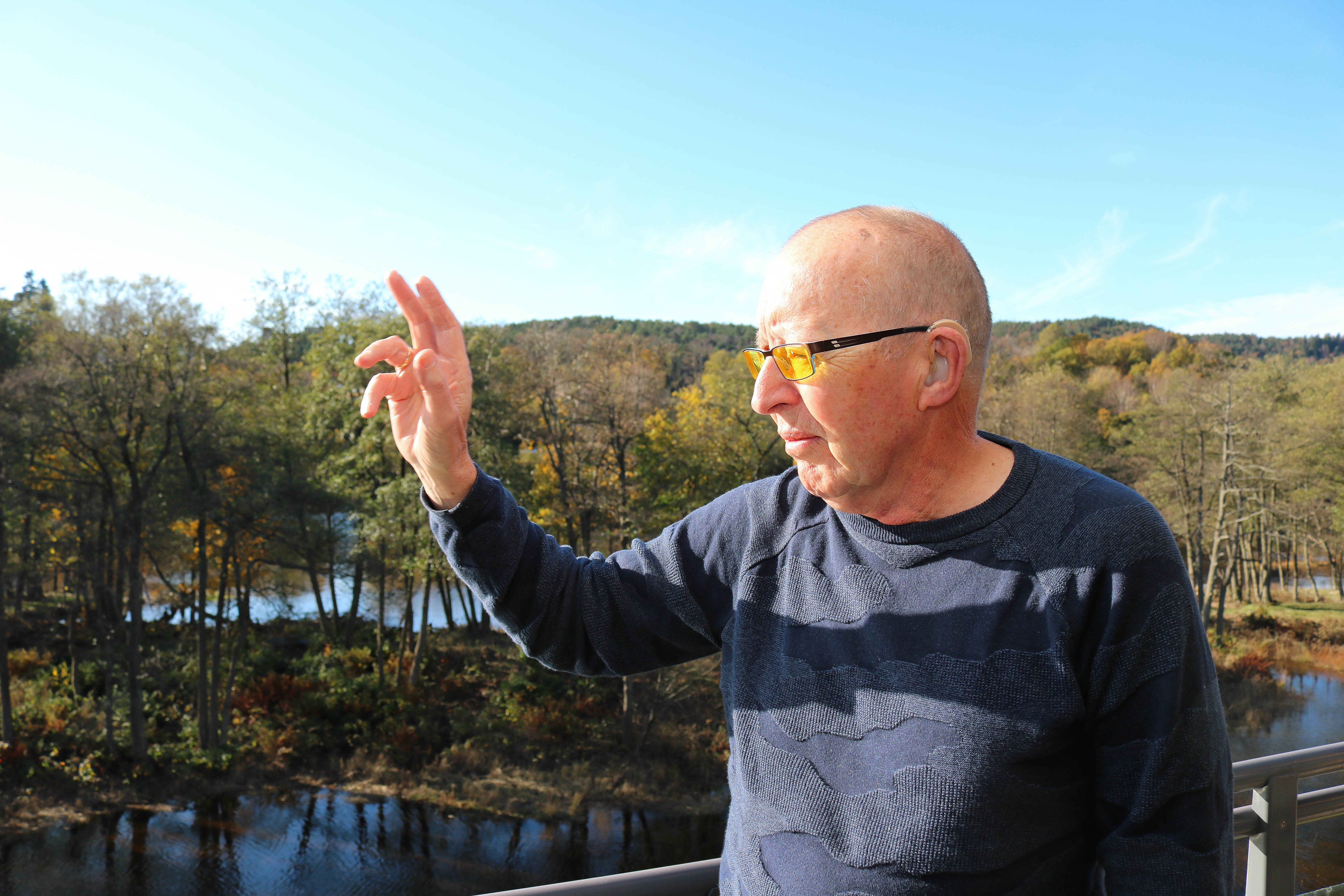 Eldre døvblind mann med gule fargefilterbriller står på sin veranda og bruker sin høyre hånd til å vise retningen til Kristiansand sentrum.