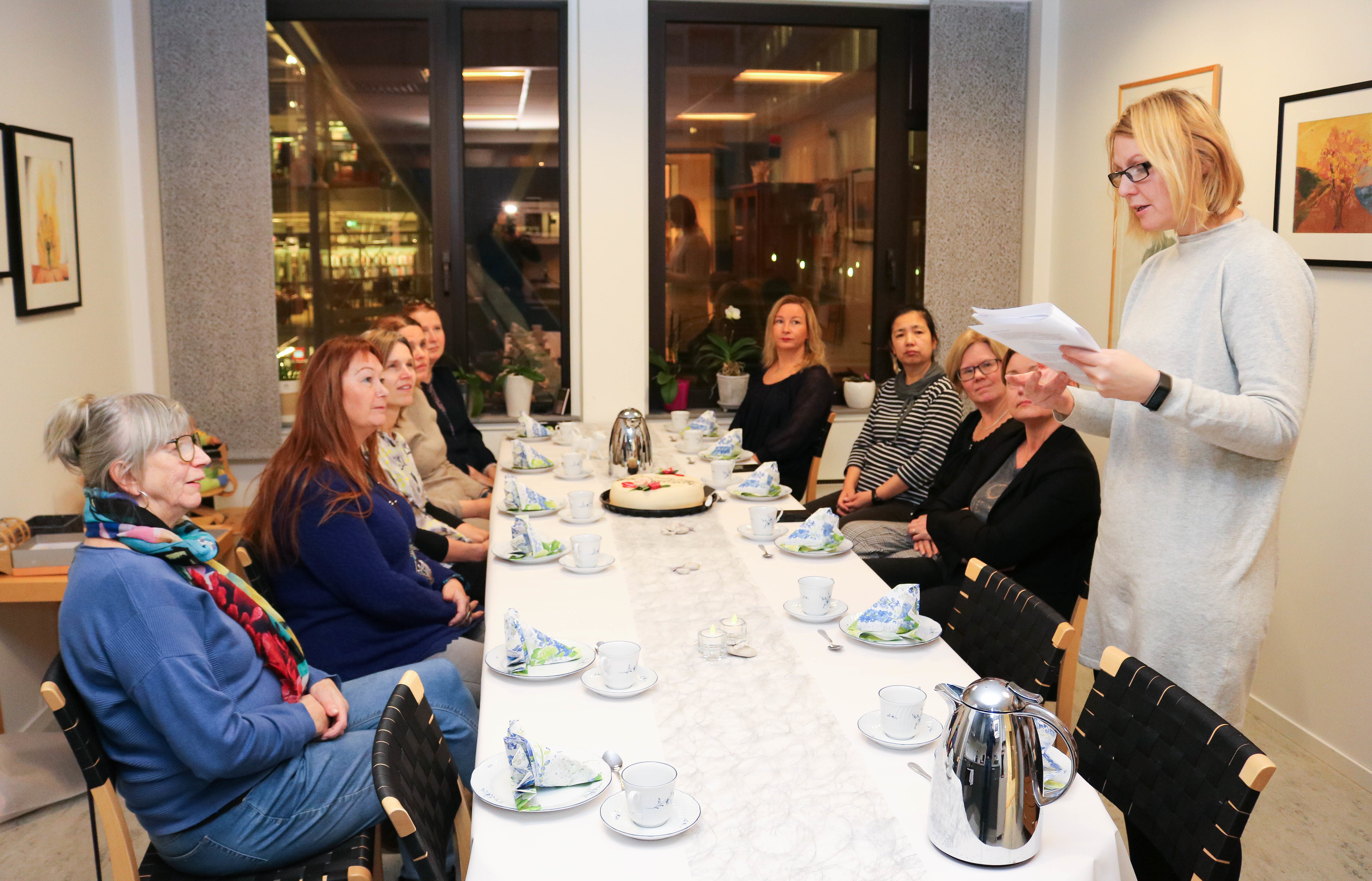 10 kvinner er samlet på et pauserom til kaffe og marsipankake, i anledning lanseringen av en ny prosjektrapport. Deres leder står oppreist til venstre og takker prosjektdeltakerne for vel utført arbeid.