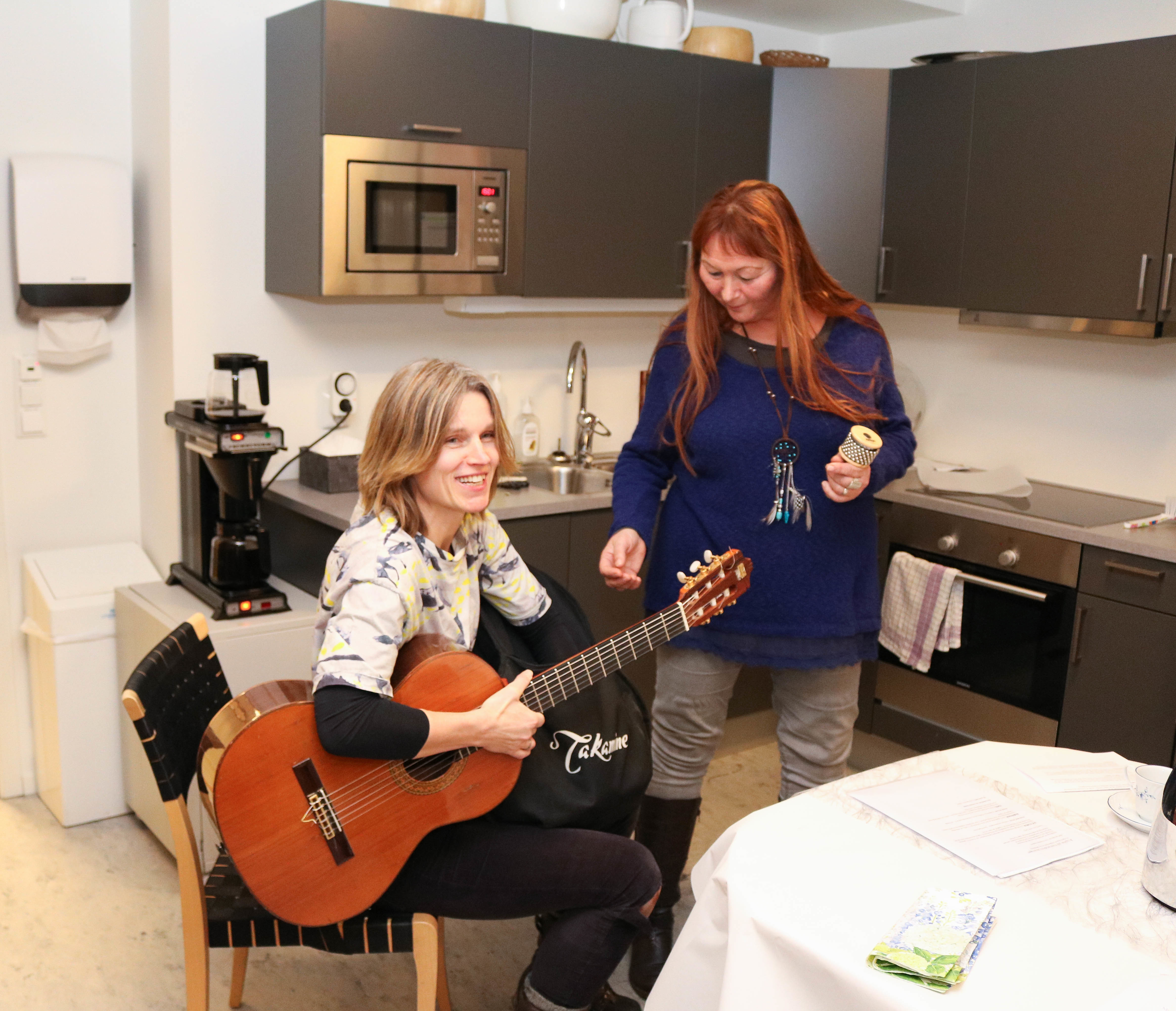 To kvinner spiller og synger sammen. Hun til venstre sitter og spiller på en gitar mens hun synger, mens hun til høyre står og synger.