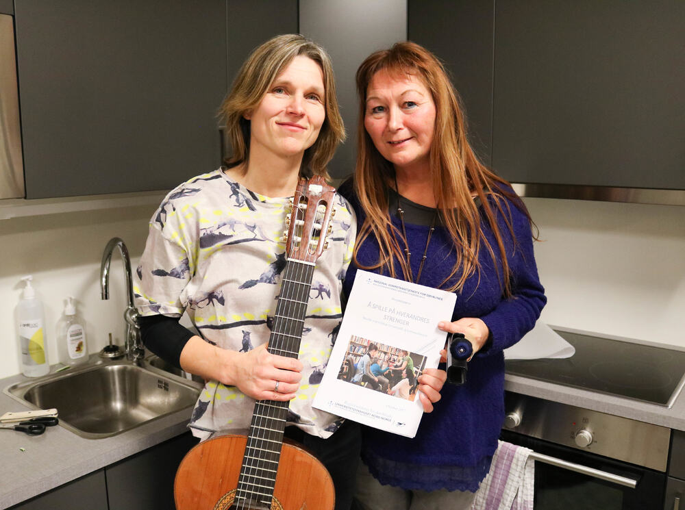 To kvinner står på et kjøkken og viser fram en prosjektrapport. Damen til venstre har lys bluse og holder en gitar, mens hun til venstre har på seg en lilla genser.
