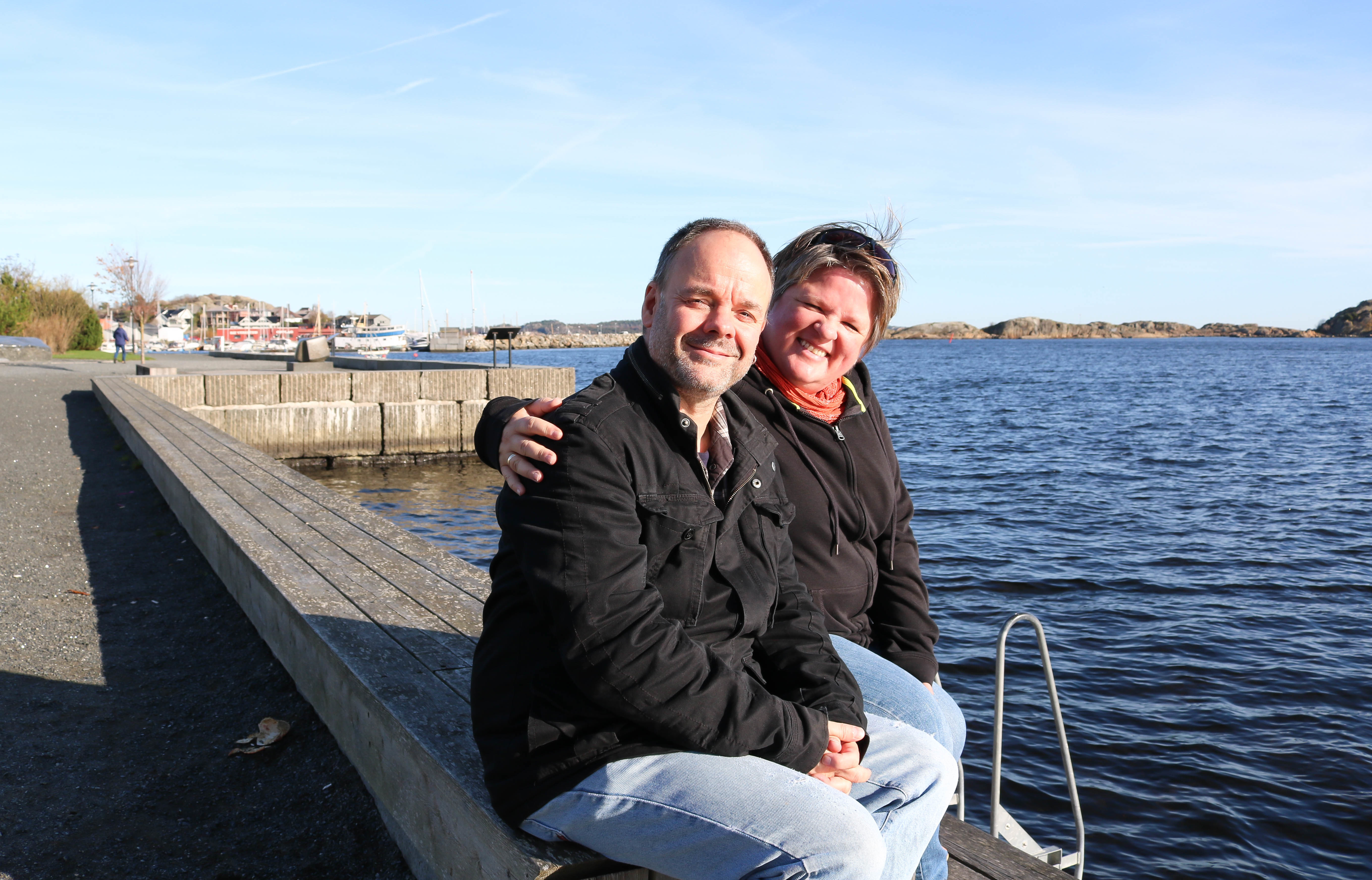 Døvblind kvinne sitter med sin ektemann nede ved sjøen på havnepromenaden i Stavern. Himmelen er blå og sol skinner på ansiktene deres. Hun holder rundt ham og de smiler. 