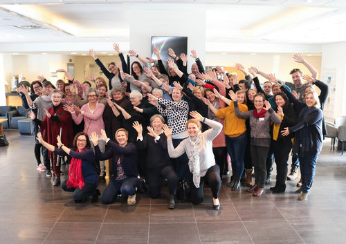Gruppebilde fra felles rådgiversamling på Asker Scandic. Foto: Roy-Morten Østerbøl. Gruppebilde av nærmere femti smilende rådgivere fra NKDBs ulike enheter. Alle strekker armene sine til høyre, som en bølge eller vifte.