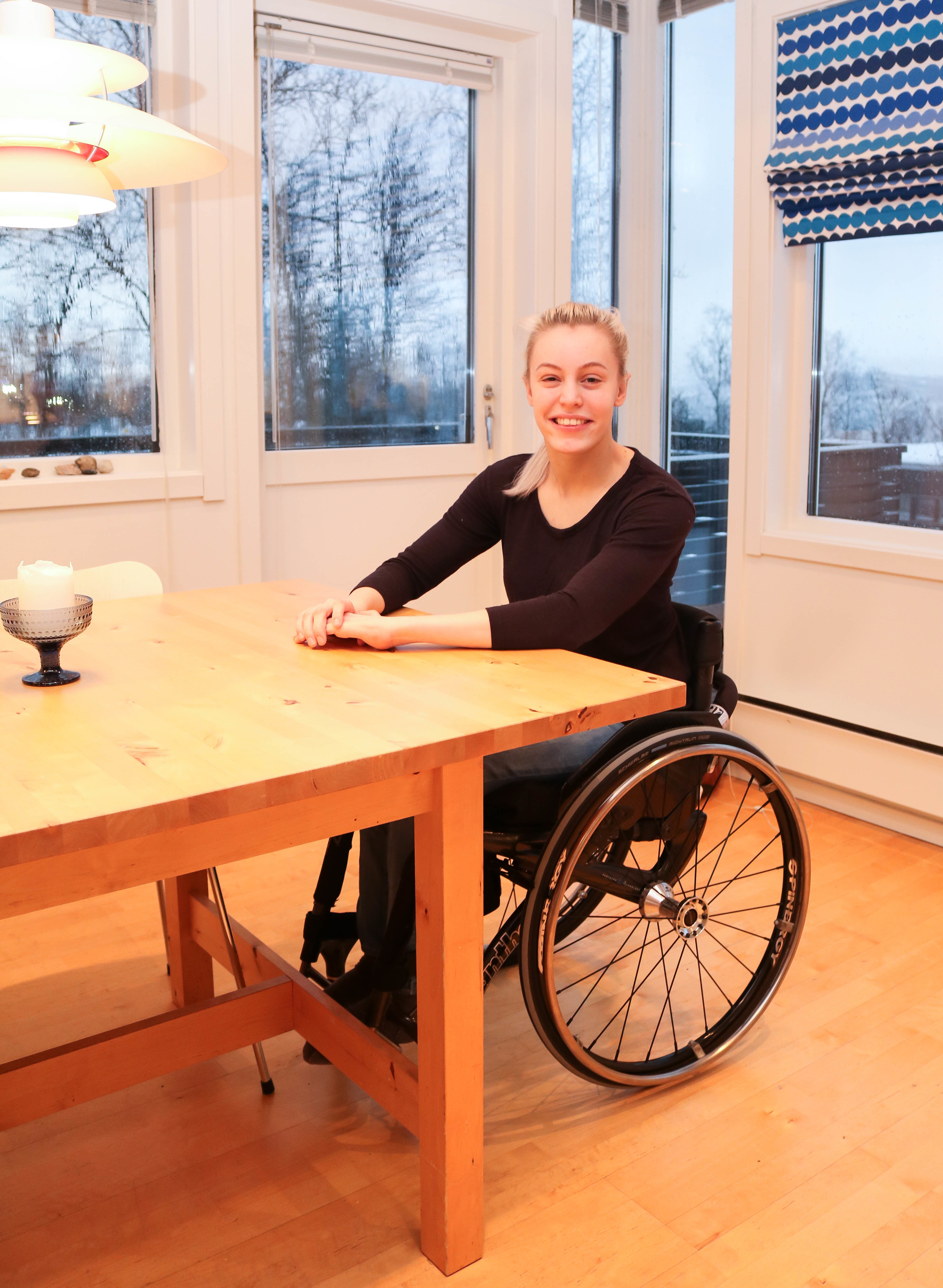 Ung smilende kvinne sitter i rullestol i enden av et stort, trehvitt kjøkkenbord.