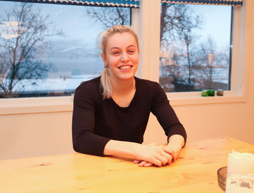 Regine Elvevold hjemme hos foreldrene sine i Tromsø. Foto: Roy-Morten Østerbøl. Ung smilende kvinne sitter med foldede hender ved enden av et kjøkkenbord.