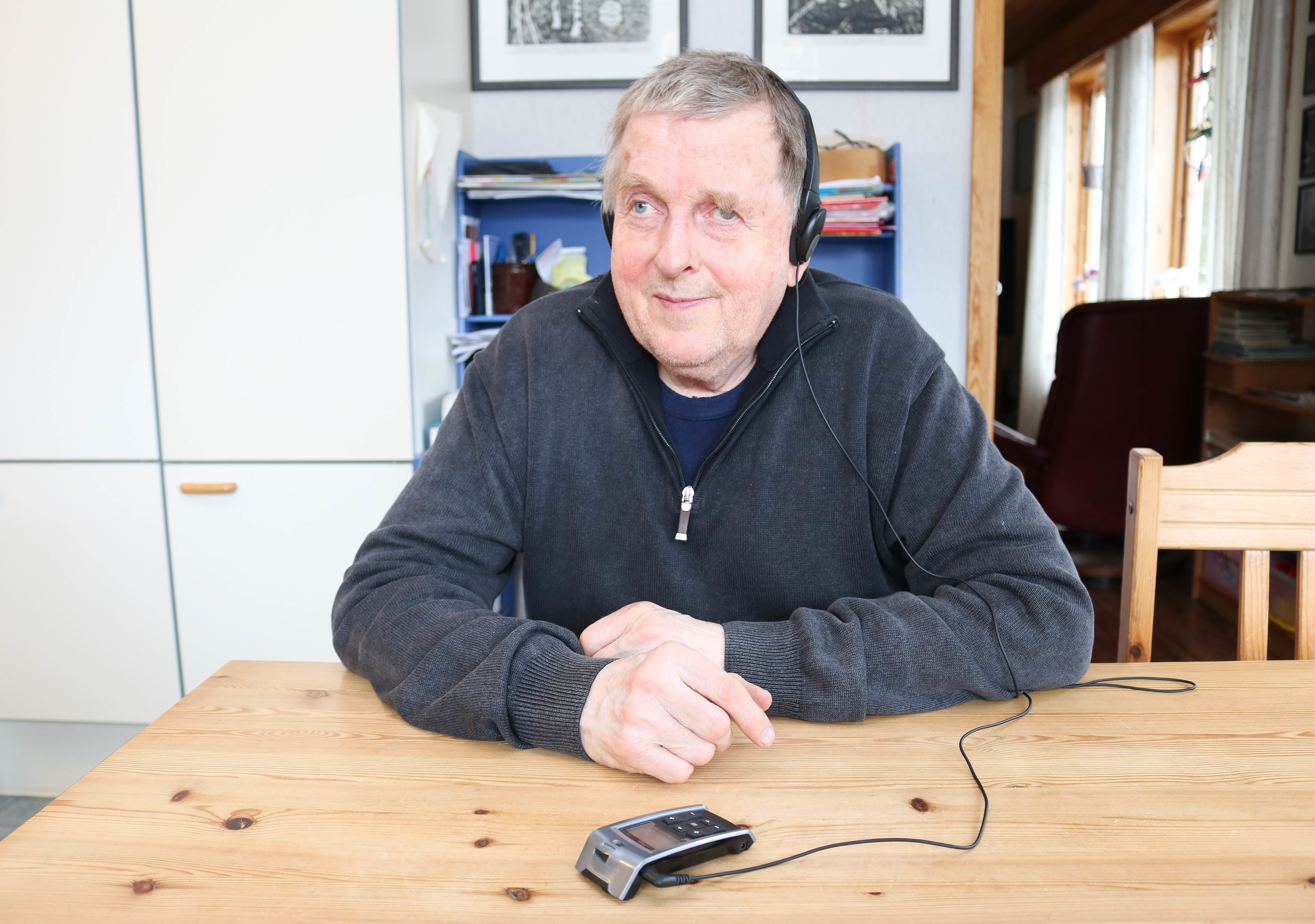 Eldre mann med grått hår og headsett sitter ved et spisbord. Foran seg på bordet har han liggende en liten taleforsterker.