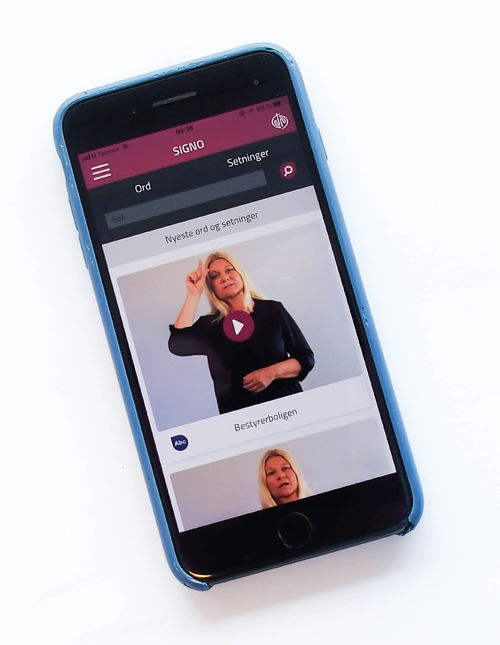 Iphone med Signos tegnspråk-app. På skjermen vises en kvinne som viser tegnet for bestyrerbolig.