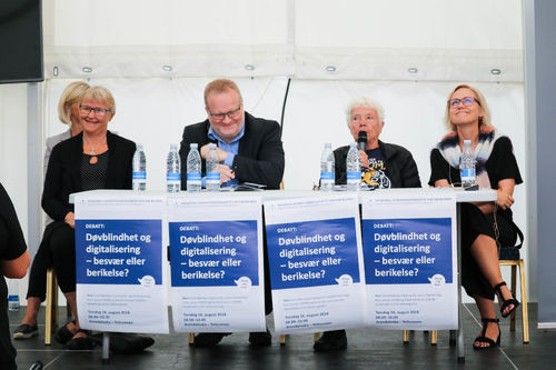 Debattanter: Åshild Johansen, Rune Jensen, Astrid Nøklebye Heiberg og Ingvild Kjerkol. Foto av Roy-Morten Østerbøl. En mann og tre kvinner i en panel-diskusjon. En rekke med plakater henger foran dem og har overskriften: Døvblindhet og digitalisering, besvær eller berikelse?
