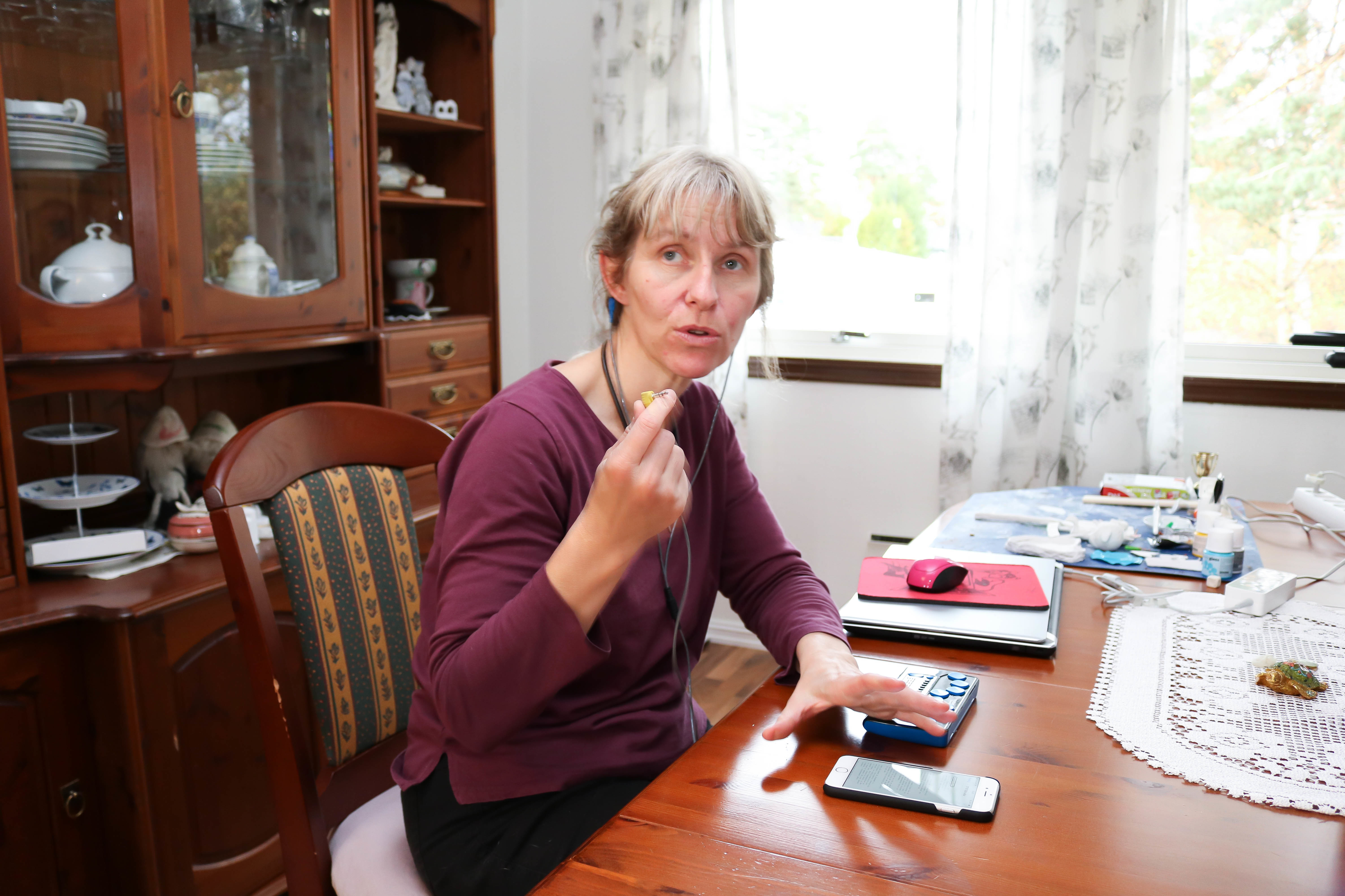 Kvinne med lilla genser i spisestue. Hun bruker sin mobil ved hjelp av headset og leselist, og holder høyrehånd mot kamera.
