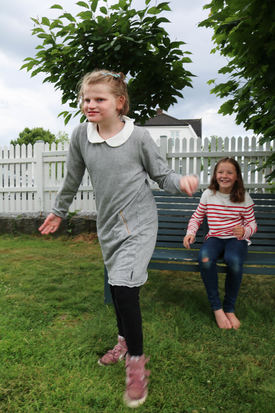 Roy-Morten Østerbøl To søstre i en sommerhage. Ei har rød og hvit-stripete genser og sitter på en benk, mens den andre går fra benken har på seg grå kjole.