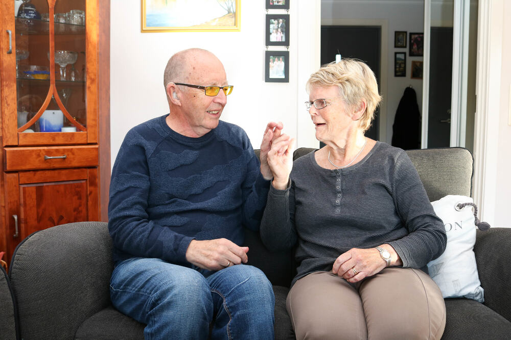 Eldre ektepar sitter tett i en sofa, de kommuniserer med taktilt tegnspråk. Mann til venstre, kvinne til høyre.