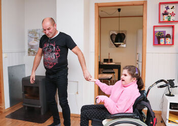 Mann ledsager sin datter i rullestol gjennom stue.