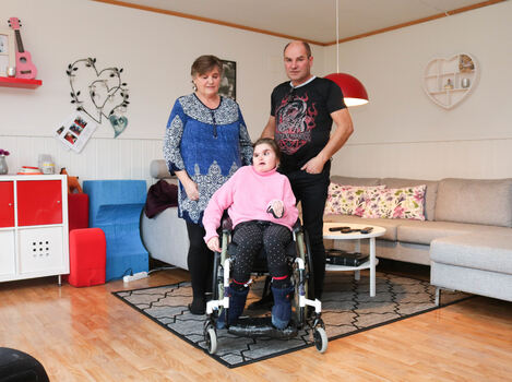 Mor og far står bak deres datter i rullestol, hun har CHARGE syndrom og de er i hennes egen stue.