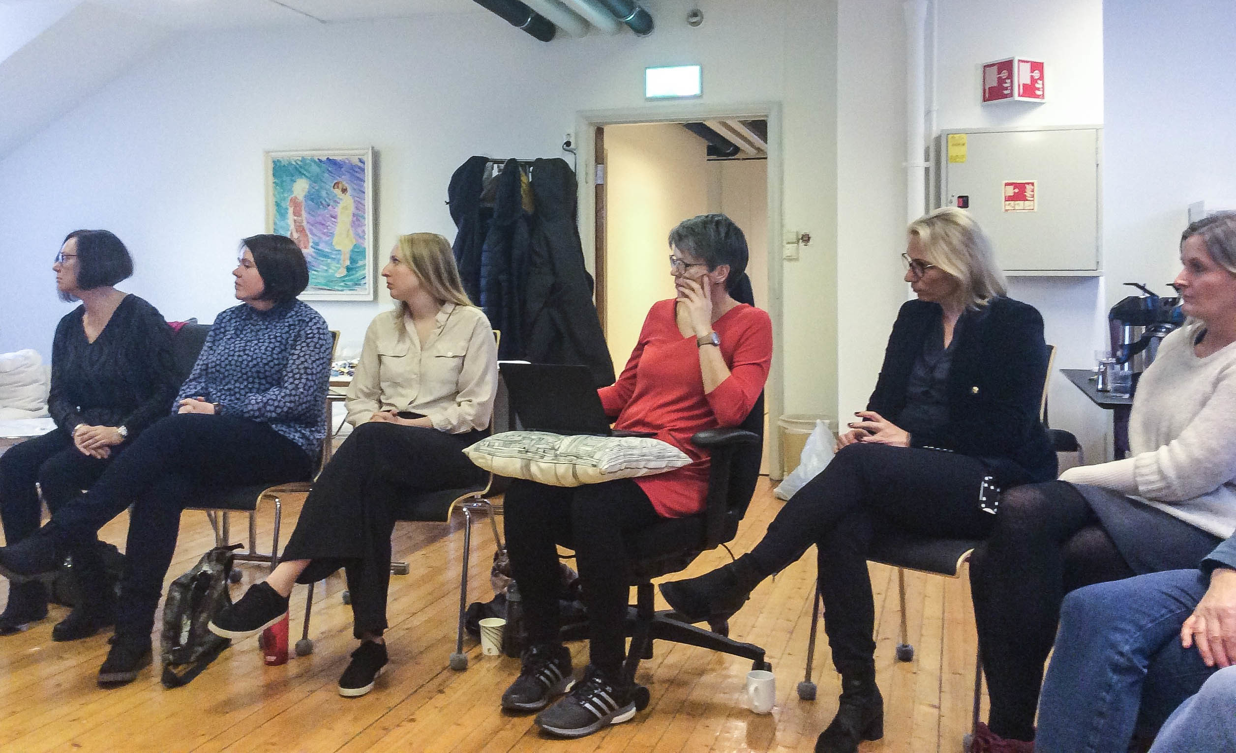 Seks kvinner sitter i en halvmåne etter hverandre i et møterom. De ser alle mot venstre i bildet.