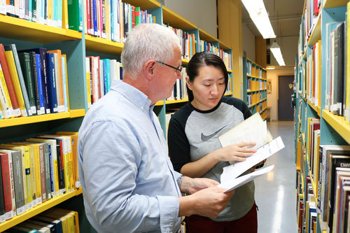Illustrasjonsfoto: Roy-Morten Østerbøl. Døvblind mann blar i en bok, sammen med en kvinnelig ledsager, mellom en rekke med gule bokreoler, inne på et bibliotek.