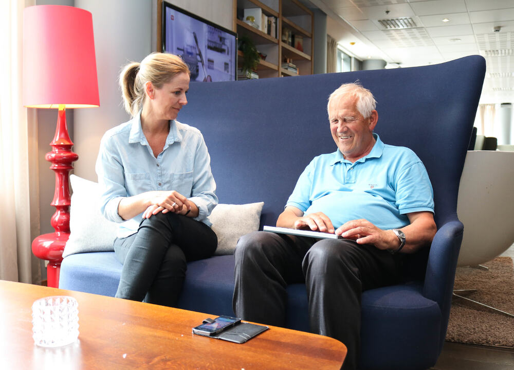 Eldre døvblind mann sitter i en blå sofa med sin ipad. En kvinnelig tolk/ledsager sitter til venstre for ham.