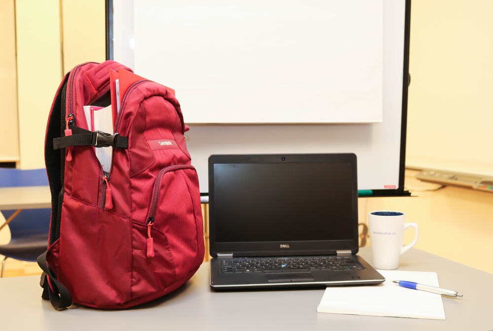 Klasserom. Rød ryggsekk med bøker som står til venstre på et bord. Ellers på border finner vi en pc, kaffekopp, penn og papir.
