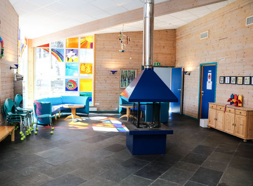 Fellesrom på skole med åpen peis midt i rommet, sollys strømmer inn fra stort, fargerikt hjørnevindu til venstre i rommet.
