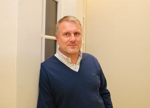 Foto: Roy-Morten Østerbøl. Smilende mann i blå genser lener seg mot en hvit vinduskarm.