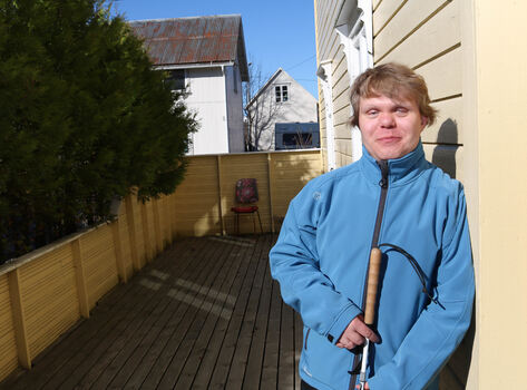 Portrettbilde av døvblind mann ved gul husvegg. Han har på seg en lyseblå fleecejakke og holder en blindestokk mot sin mage.