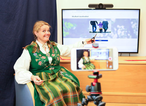 Koordineringsenhetens leder Trine Seljeseth åpner NKDBs nye nettkurs om medfødt døvblindhet. Foto: Roy-Morten Østerbøl. Kvinne i bunad klipper lanseringsbånd foran tv med nettkurs, i forgrunnen mobil på stativ som filmer det hele.