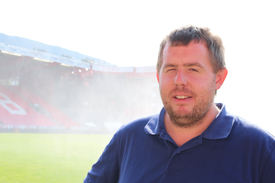 Roy-Morten Østerbøl Mann i blå piquet poserer inne på Brann stadion med vannsøyle i bakgrunnen.