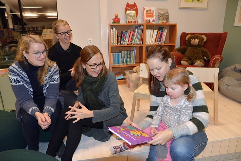Fra venstre: helsesykepleier Renate Sætertrø, leder helsestasjons- og skolehelsetjenesten Barbro Orskaug, biblioteksjef Katrine Herringbotn, Astrid Jemblie Aga og datteren Sigrun. Bilde av mor og barn sammen med ansatte ved helsestasjons- og skolehelsetjenesten som leser bok