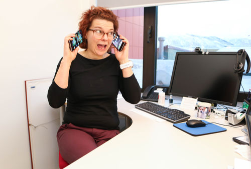 Kvinne ved arbeidspult holder oppe to mobiltelefoner, en mot hvert øre.