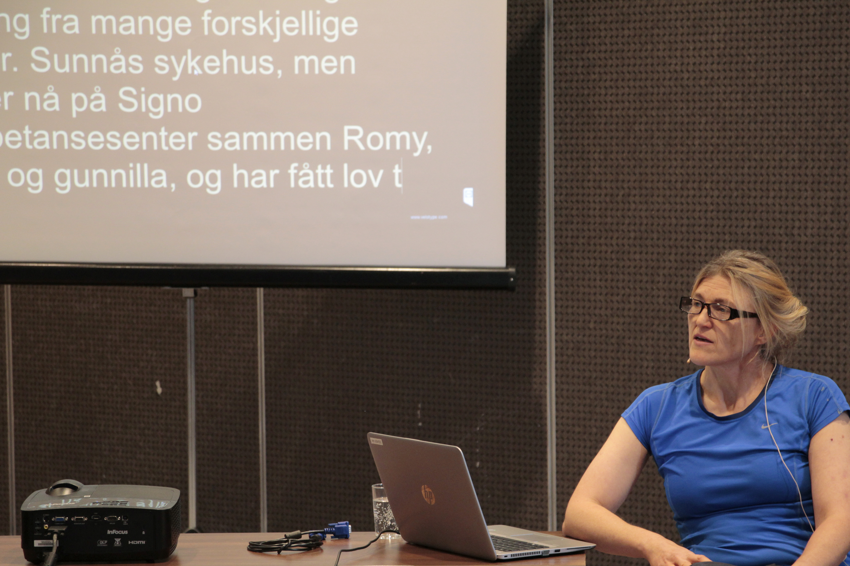 Kvinne holder foredrag sittende ved sin laptop, i bakgrunnen en overhead med diverse tekst. 