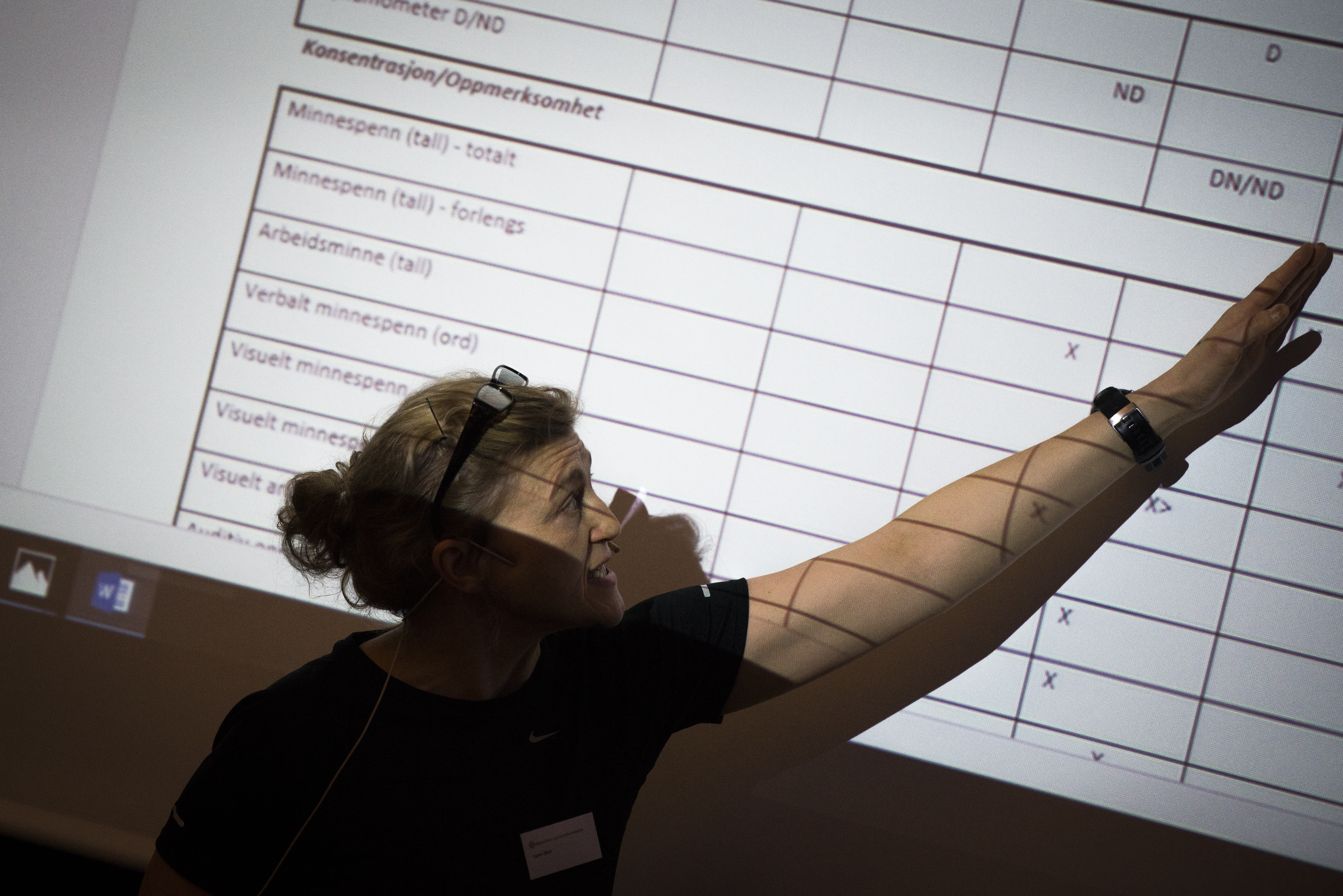 Kvinne i forelesningssituasjon, hun peker med venstrearm på detaljer i en opplyst og forstørret tabell. 