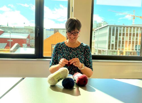 Administrasjonskonsulent Eva Hirsti har tatt oppfordring til DbI og er i gang med å strikke lapper til et garngraffiti-verk i Tromsø. Foto: Roy-M. Østerbøl. Glad kvinne som strikker i solfylt kontorlokale.