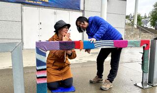 Kampanjearbeid: Administrasjonskonsulent Eva Hirsti og renholder Sun Jie monterer sammensydde strikkelapper på et sykkelstativ ved Tromsø rådhus. Foto: Roy-M. Østerbøl. To kvinner jobber med sette i stand garngraffiti på et metallrekkverk.
