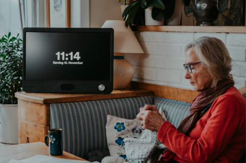 Eldre kvinne med strikketøy sitter i sofaen foran en stor skjerm, som bare har en knapp. På skjermen står det 11:14 søndag 15. november.