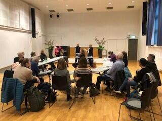 Foto: Statped. En gruppe folk sitter rundt en sirkel med møtebord, fremst står to personer og kommuniserer via tegnspråk.
