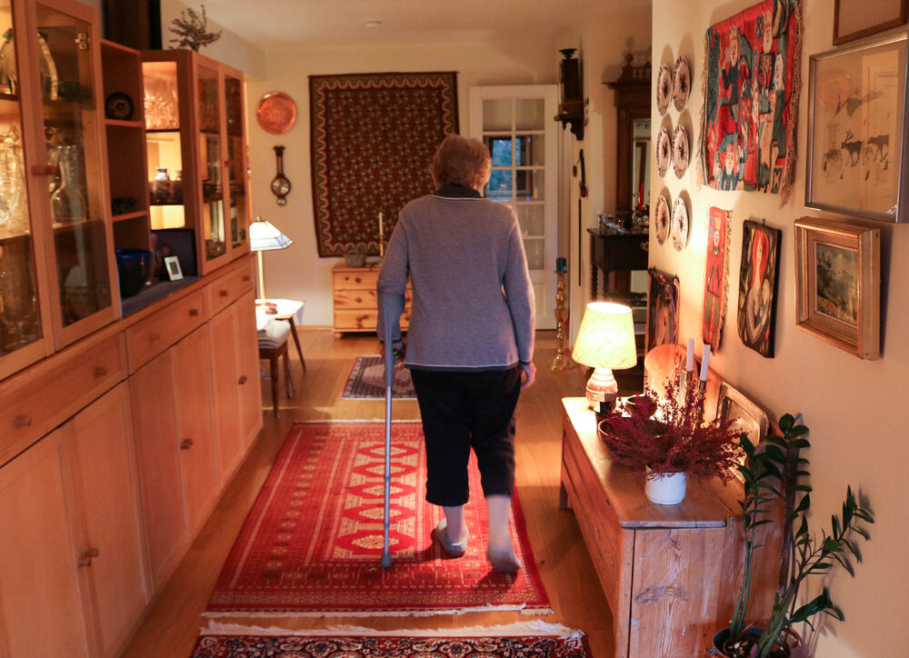 Eldre kvinne går gjennom sin stue med en krykke i venstre arm.