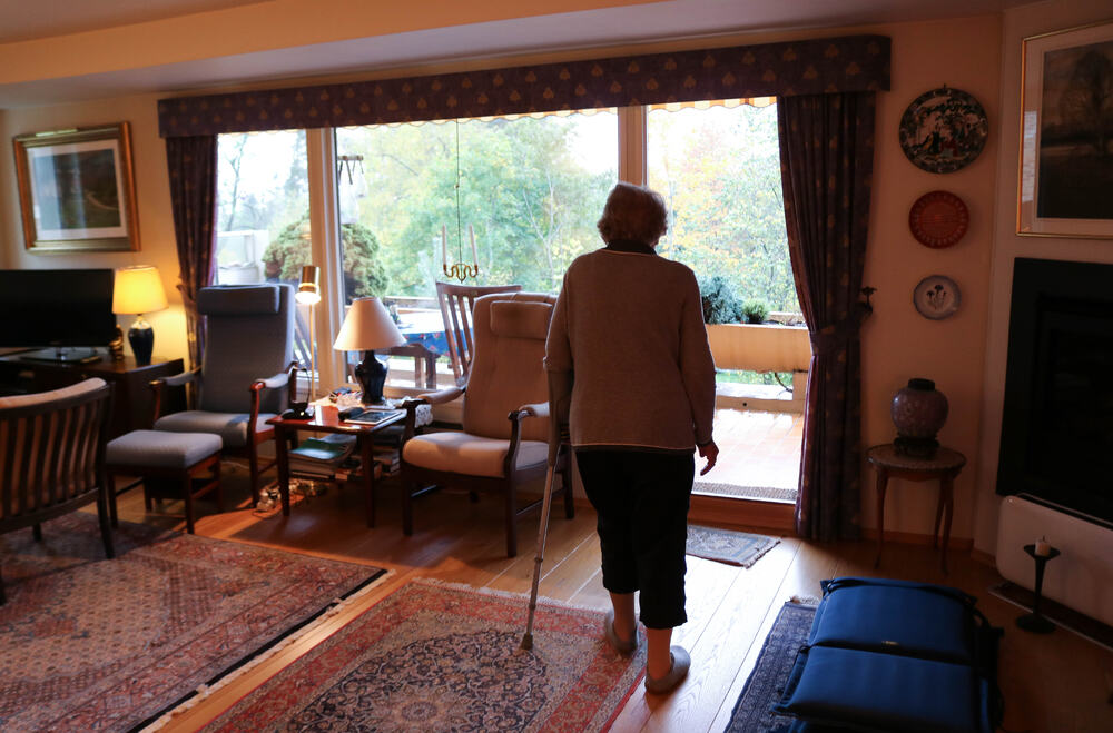 Eldre kvinne går gjennom sin stue med en krykke i venstre arm.
