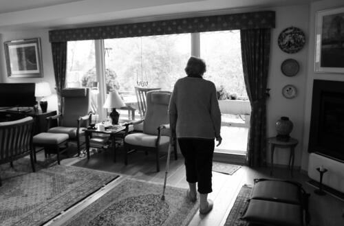 Eldre kvinne går gjennom sin stue med en krykke i venstre arm. Sort-hvit versjon.
