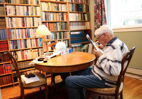Eldre mann leser et magasin med hjelp av lupe inne på sitt personlige bibliotek.