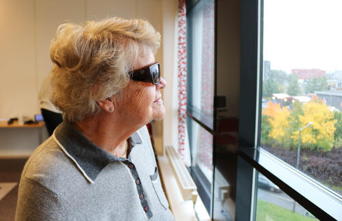 Eldre kvinne med oransje filterbriller ser ut av et vindu.