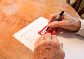 Eldre person skriver sin underskrift på et ark med hjelp av en rød plastmal.