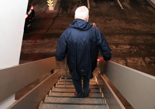 Eldre mann med blå hettejakke går ned en bratt trapp, han bruker en krykke til hjelp.