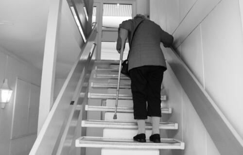 Eldre kvinne går opp en trapp med krykke i venstre arm. Sort-hvit versjon.