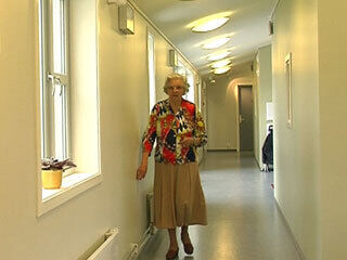 Eldre kvinne som kommer mot oss i en ryddig korridor, med god belysning.