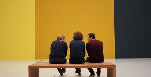 Tre personer på en benk, foran en vegg malt i sterke fargeblokker