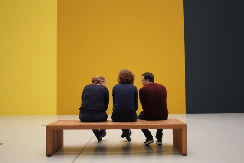 Tre personer på en benk, foran en vegg malt i sterke fargeblokker