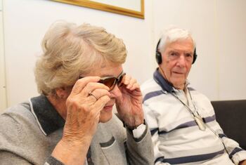 Eldre kvinne tester oransje filterbriller, henne mann sitter ved henne med headsett.