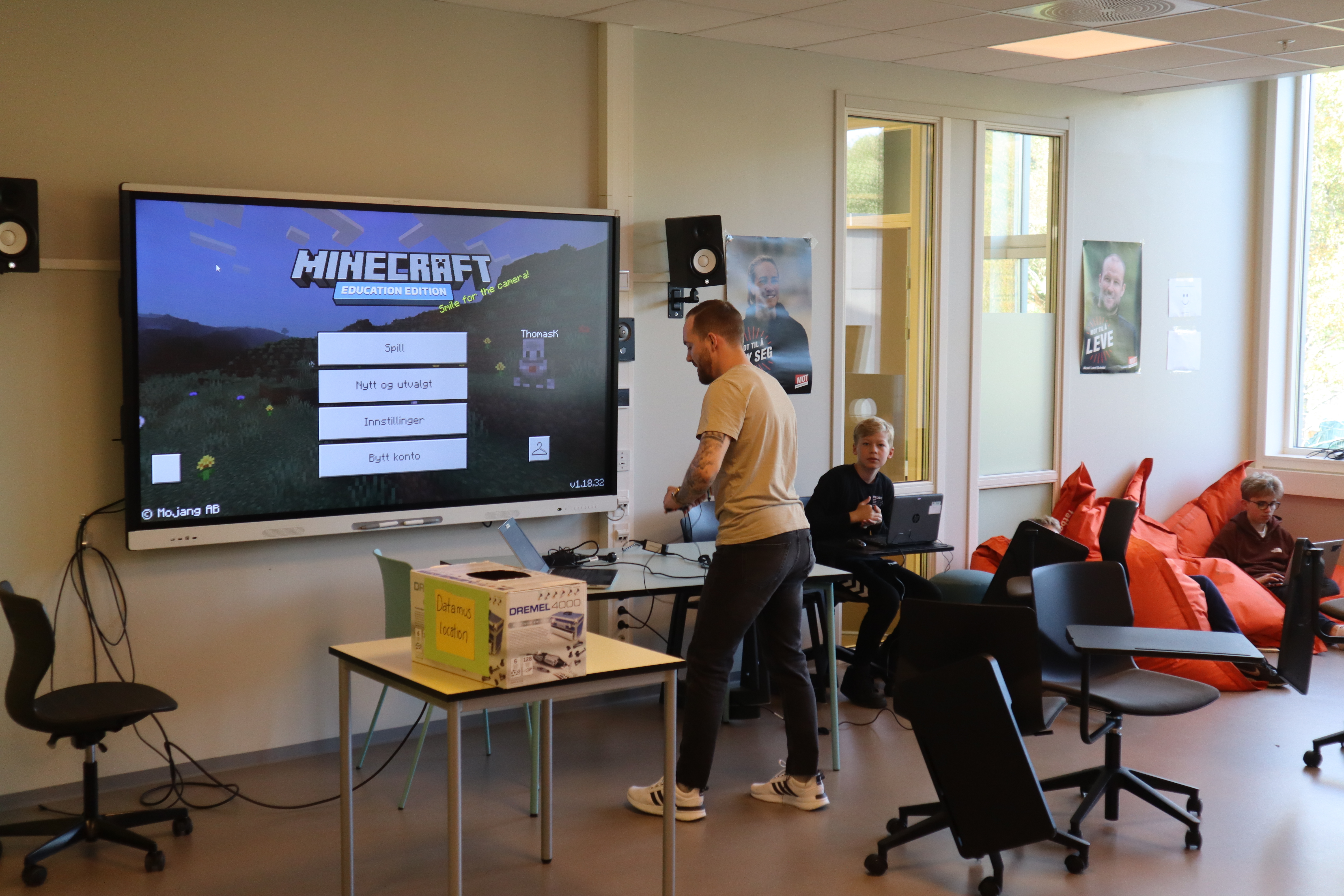 Et klasserom med en skjerm som viser spillet, lærer Thomas Kristiansen og tre elever i bakgrunnen.