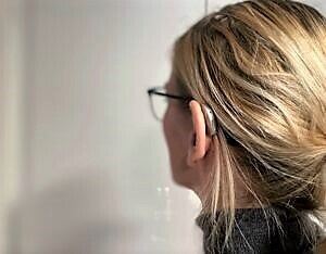 Profilfoto av kvinne med høreapparat og briller, hun ser inn i en grå vegg.